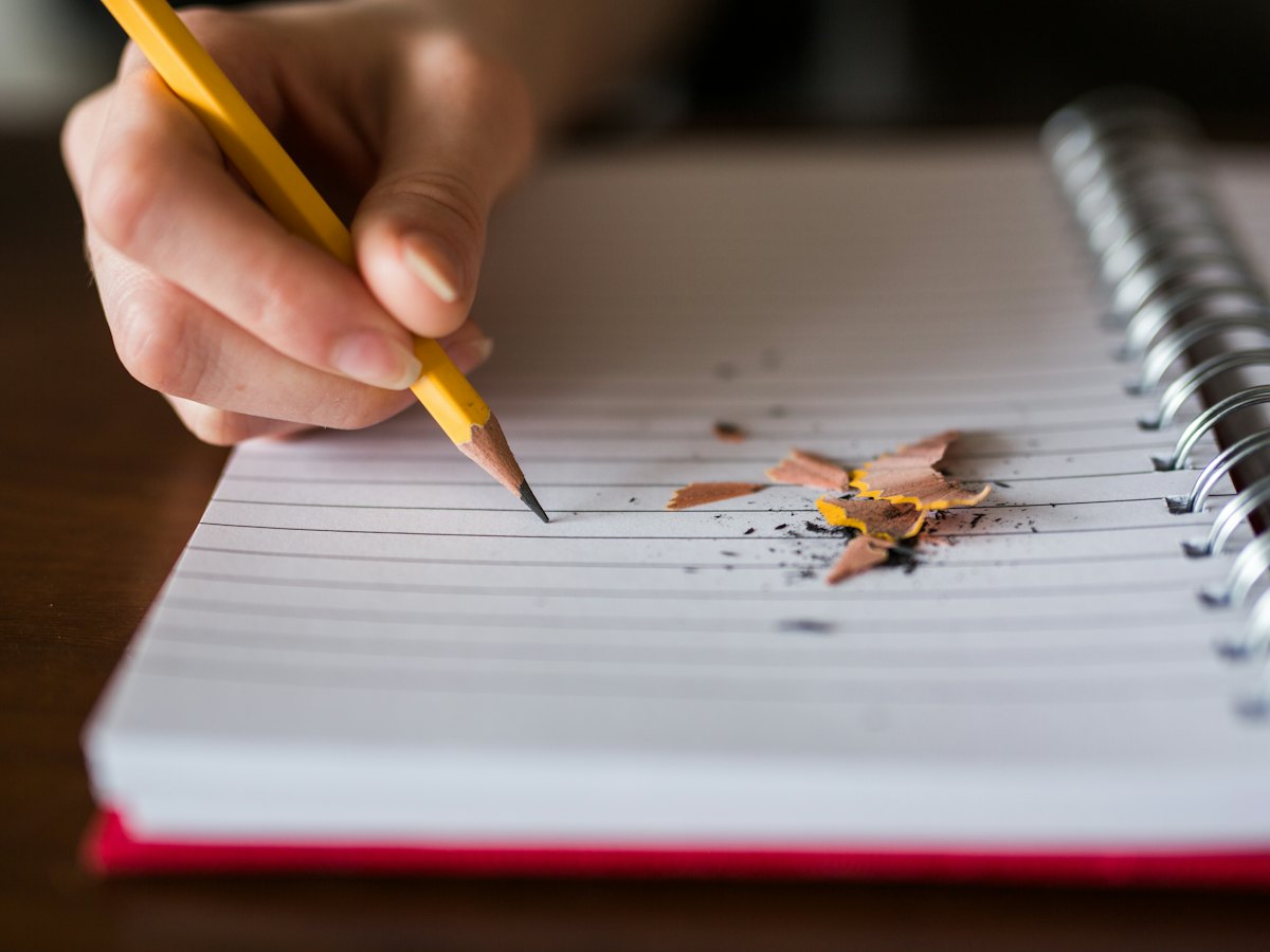 A hand holding a pencil, writing in a spiral notebook on a wooden desk