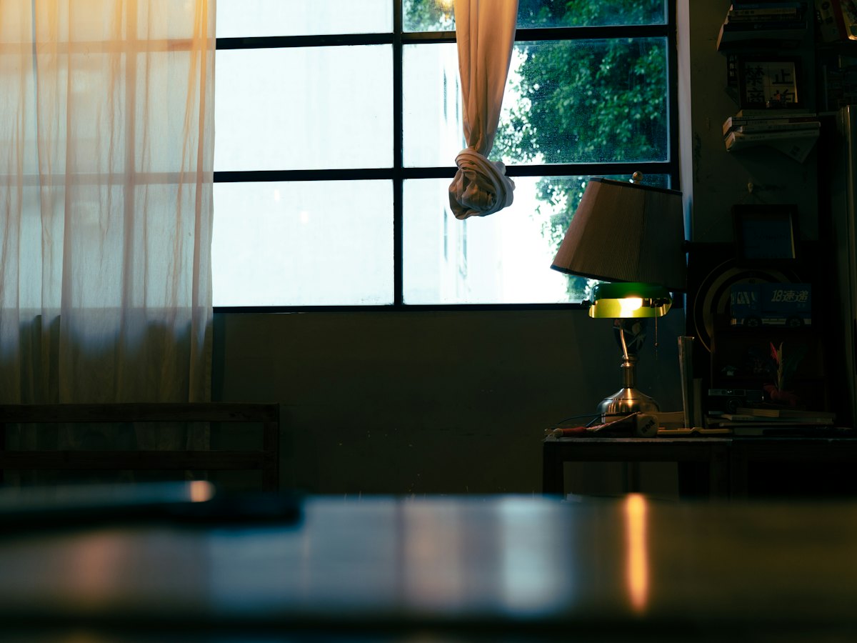 A lit desk lamp on a table at evening, warm light through a curtained window