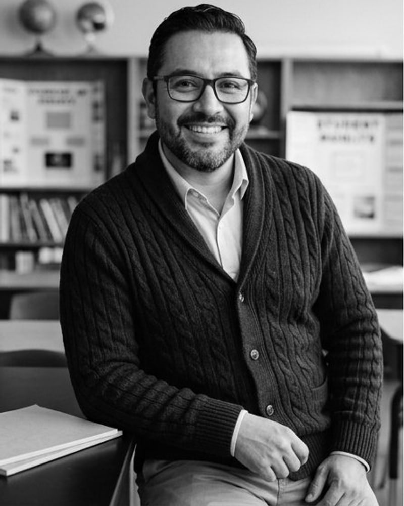 Black-and-white portrait of a smiling man with glasses in a cable-knit cardigan, seated in a classroom
