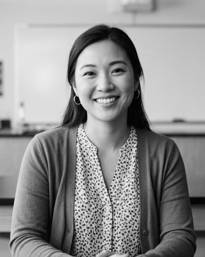 Black-and-white portrait of a smiling young woman in a cardigan, seated at a classroom table