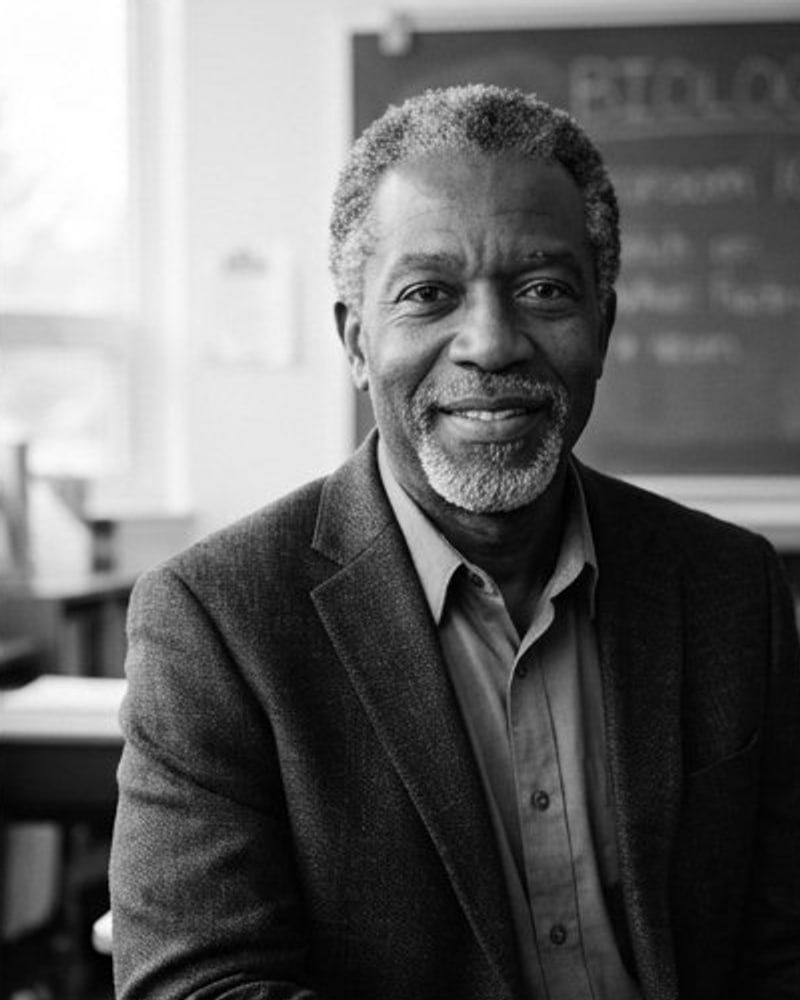 Black-and-white portrait of a smiling older man with a gray beard in a tweed jacket, seated by a chalkboard